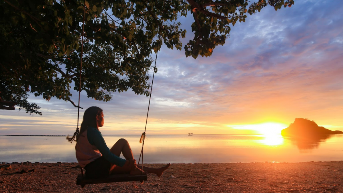 A young woman sits on a swing overlooking the setting sun over the water.