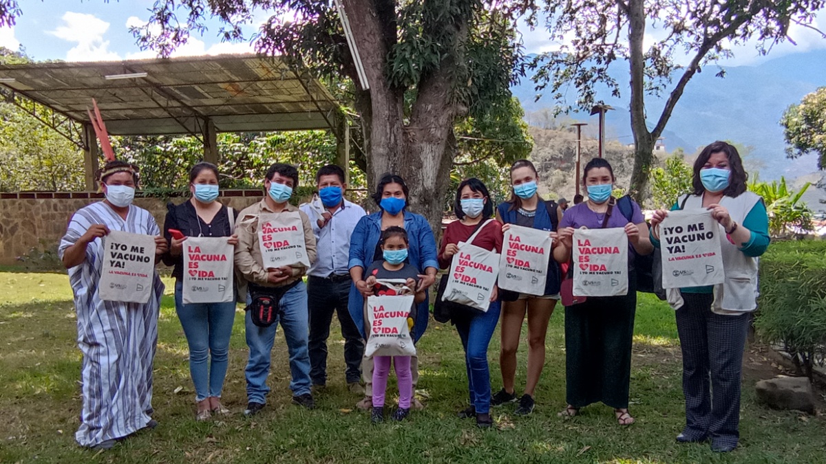 Indigenous women and men standing in the outdoors holding bags with printed messages supporting COVID 19 vaccination.