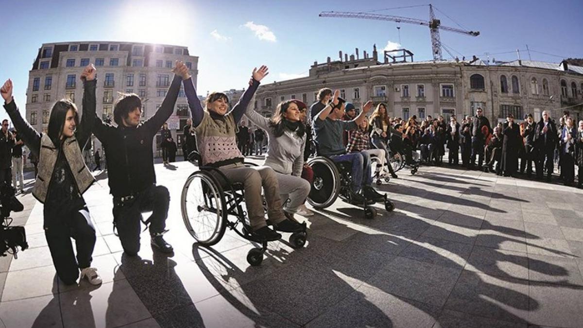 A large group of young people, including several wheelchair users, stand in line holding hands in a public square.