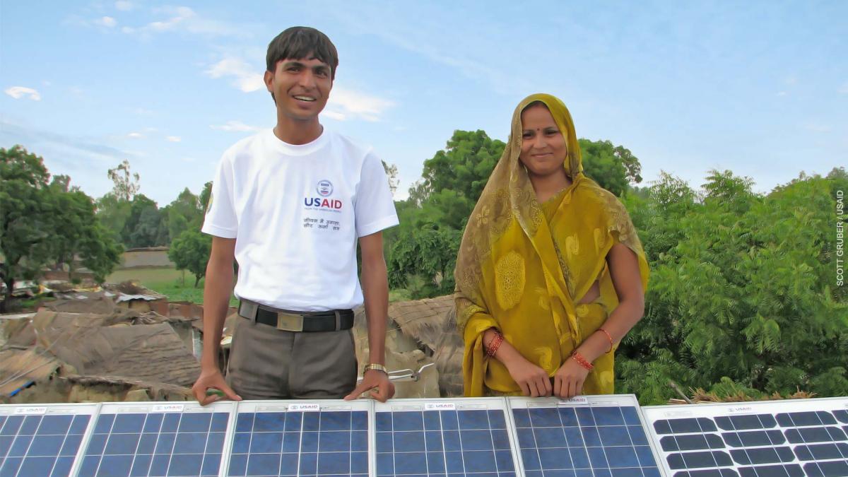 A smiling Indian man and woman stand behind a solar array in a rural village