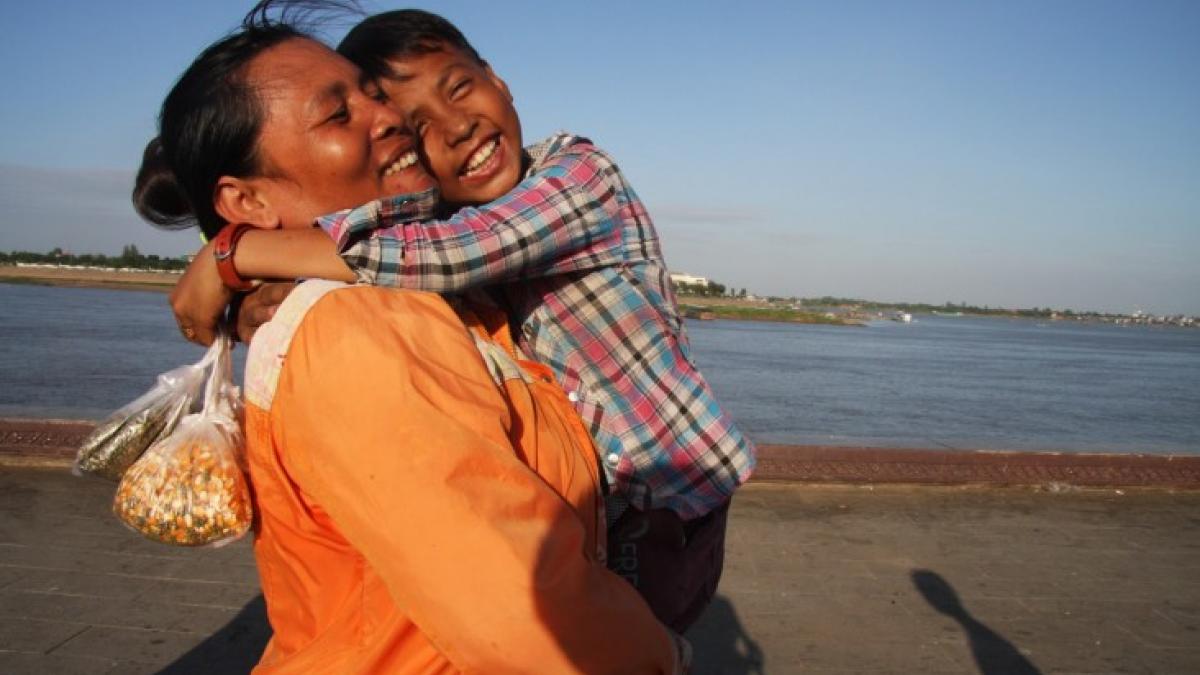 A smiling boy hugging his mother near a large body of water.