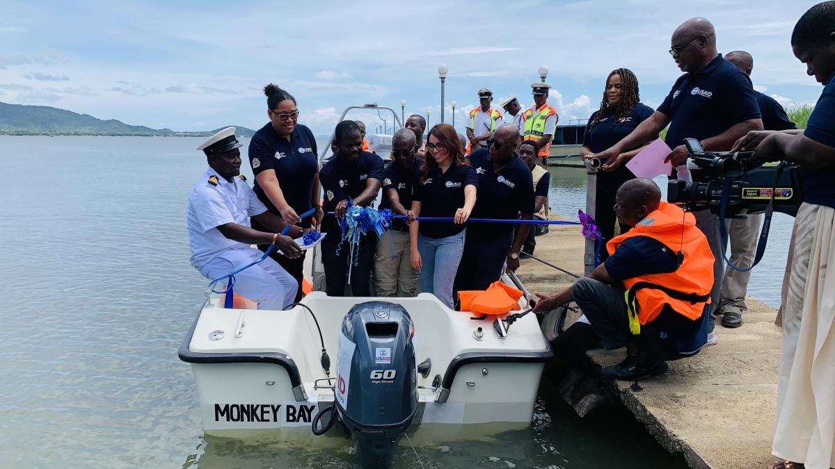 USAID and Government of Malawi officials cutting a ribbon in a boat in the presence of other district fisheries officials, law enforcers, and the media.
