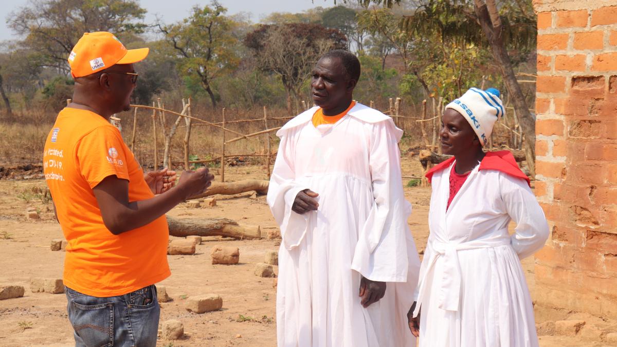 A Zambian man in a USAID t-shirt is talking to a Zambian man and woman who are dressed in the white robes of religious leaders in their church