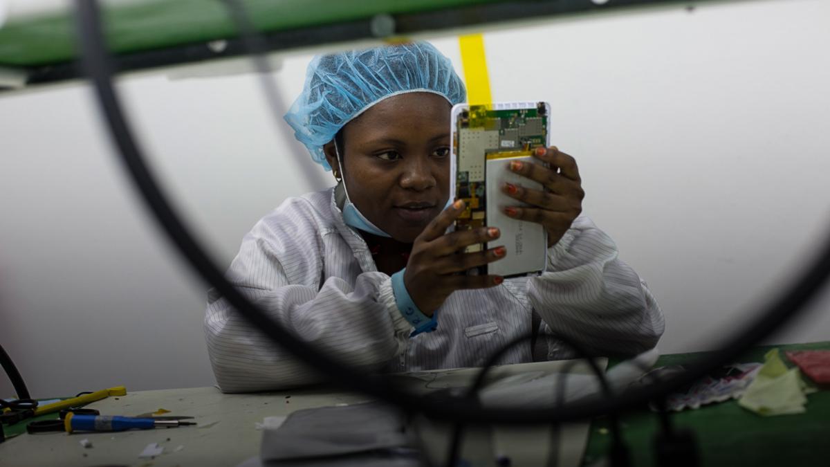 A young woman in a hair net and lab coat examines a tablet in the process of being assembled.