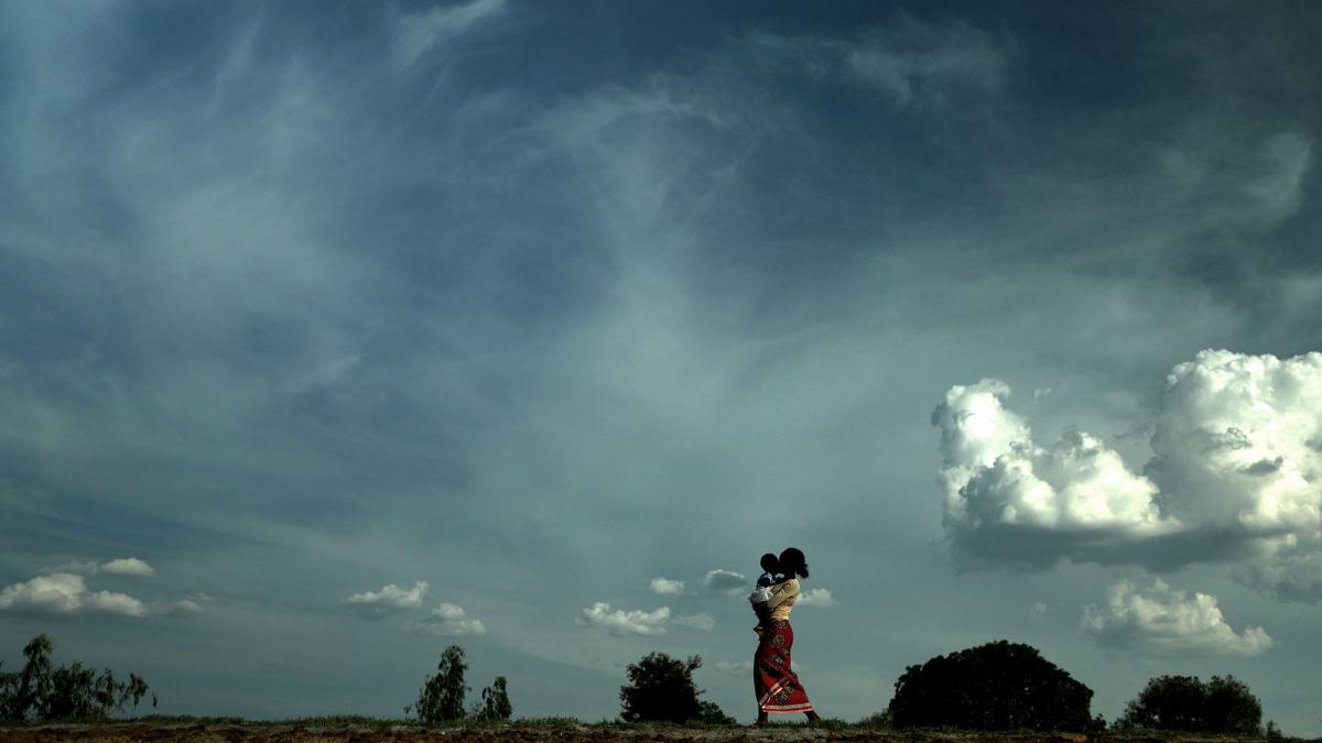 In the distance, a woman holds a baby, with sky and clouds in the background.