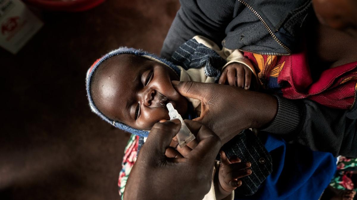 A closeup overhead shot of an infant child in Malawi held by the mother. The mother squeezes the child’s cheeks to hold its mouth open as a health worker squeezes medicine into the child’s mouth. 