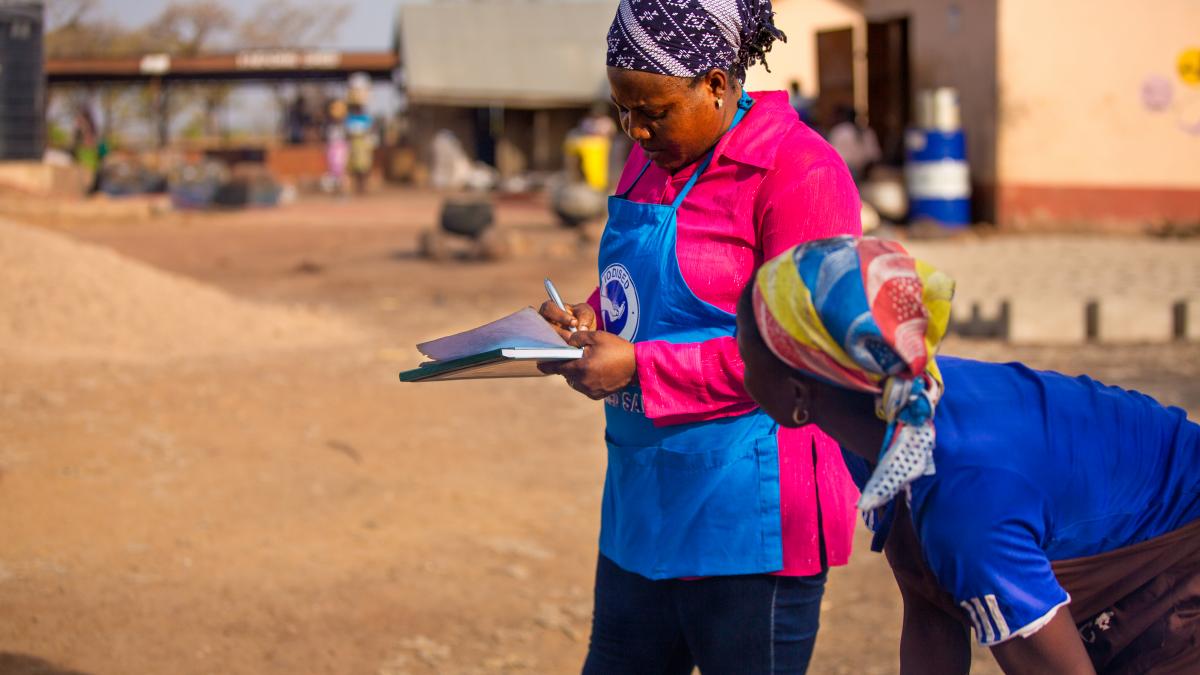 Two women stand in a village in Ghana, while one of them writes something down on a notebook she is holding.