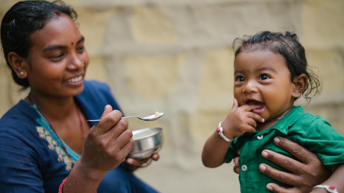 A woman and a young child smile as the woman holds a spoonful of food for the child