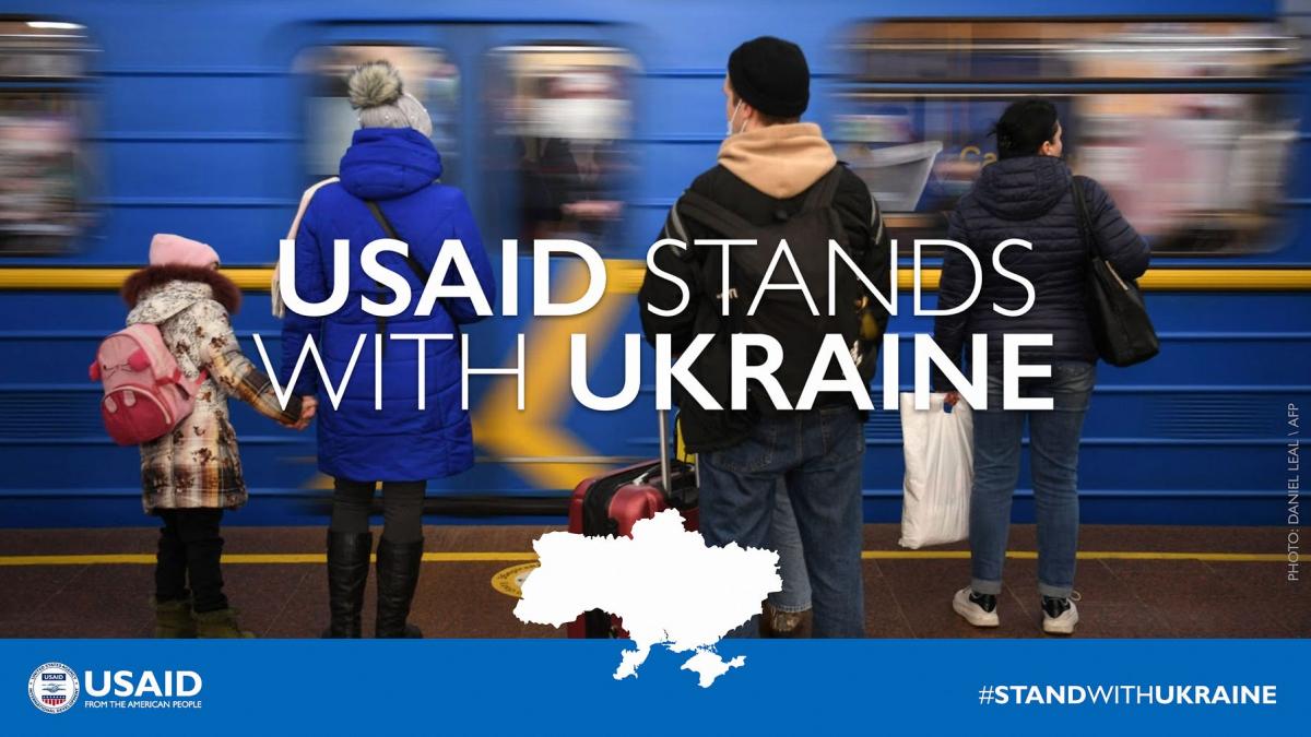 An image of 4 people standing in front of a moving blue metro train. Picture has the text "USAID Stands with Ukraine."