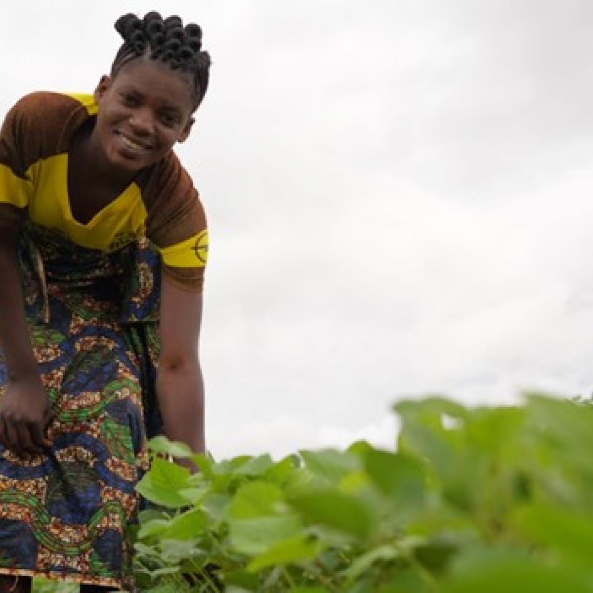 A young women smiles while kneeling down to display a field of green soybean plants