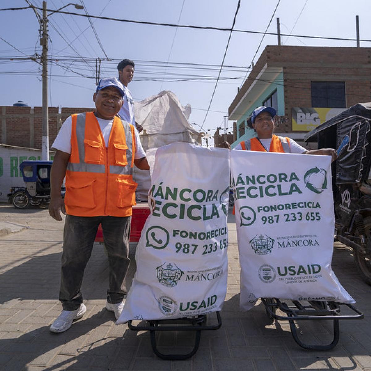 A couple of local workers holding two bags full of plastic waste. The bags have the name of the project and the USAID logo.