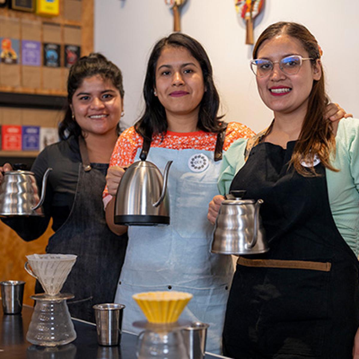 Three young barista women smiling and holding pots of brewed coffee