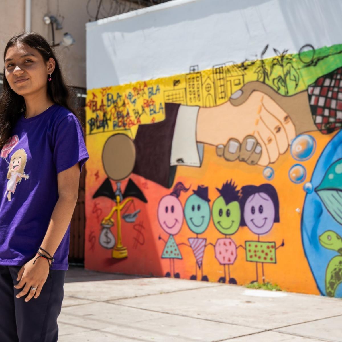 A girl in a purple shirt stands in front of a bright mural at her school.