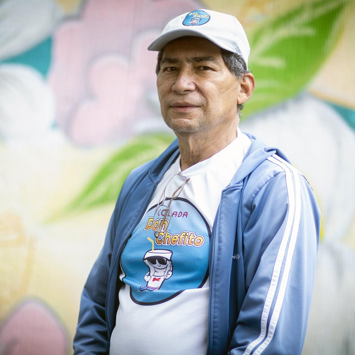 This picture shows Carlos Torres’ upper body and face. He is wearing a blue sports jacket and a hat, and is standing in front of a colorful mural with flowers.