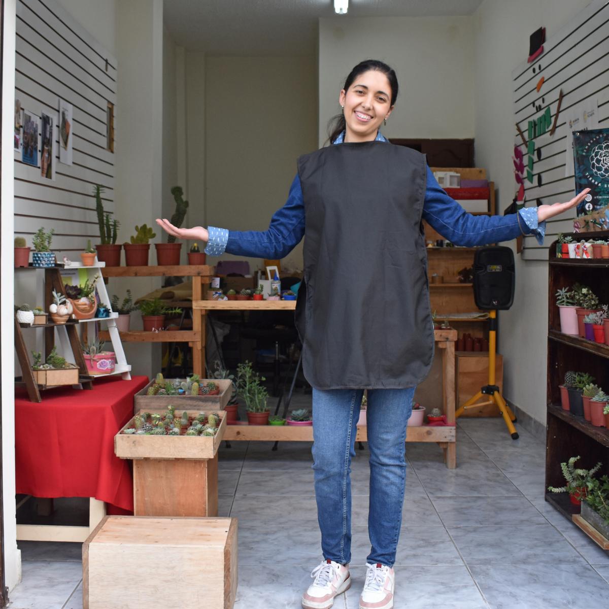 This is a picture of Omaira, the main character, standing in front of her little store with arms open, welcoming people. She is in jeans, a long-sleeved shirt, sneakers, and and black apron. There are plants on shelves.