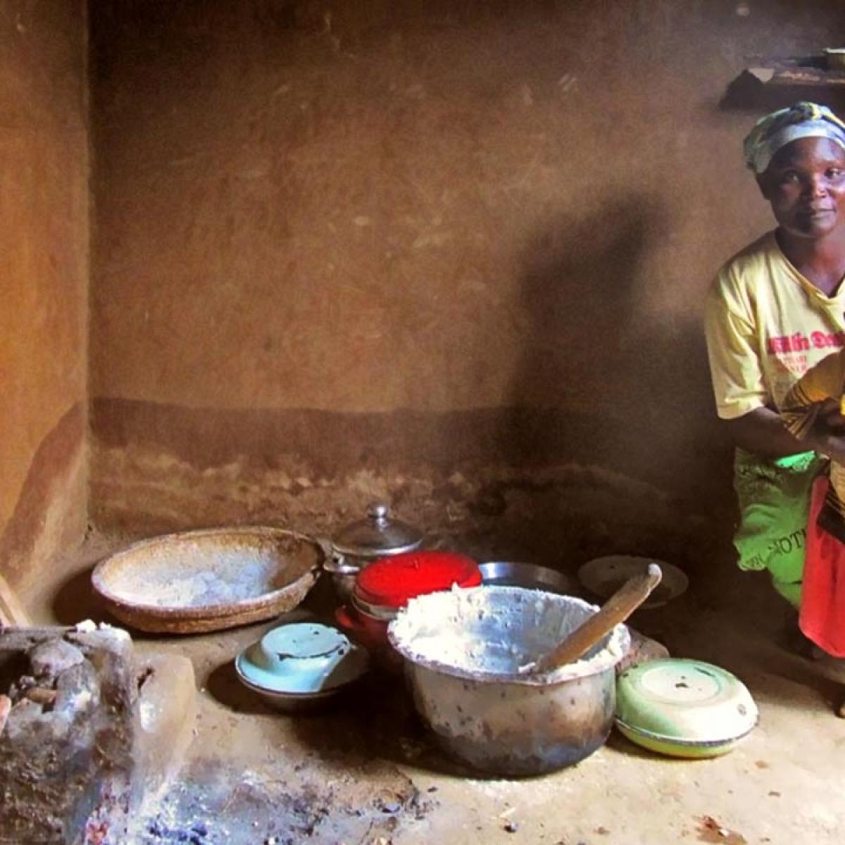 A Malawi woman and her toddler daughter in the cooking area of their small home. A small wood-burning cookstove sits on the floor near mixing bowls.