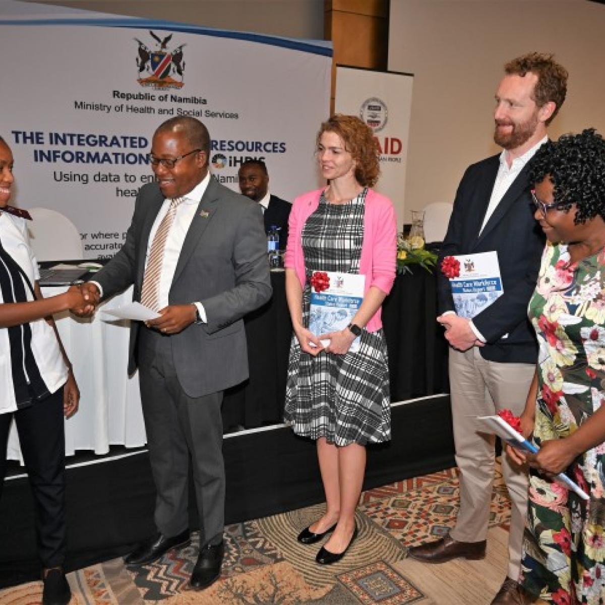 Ms. Letisia Haidula shakes hand with Mr. Ben Nangombe while Jessica Long and a couple representatives observe. The group stands in front of a table and screen about the Republic of Namibia's Ministry of Health and Social Services.