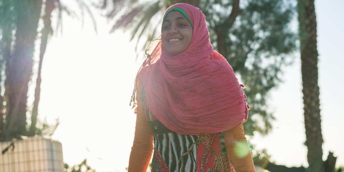Young woman wearing pink head cover smiles as she stands outdoors.