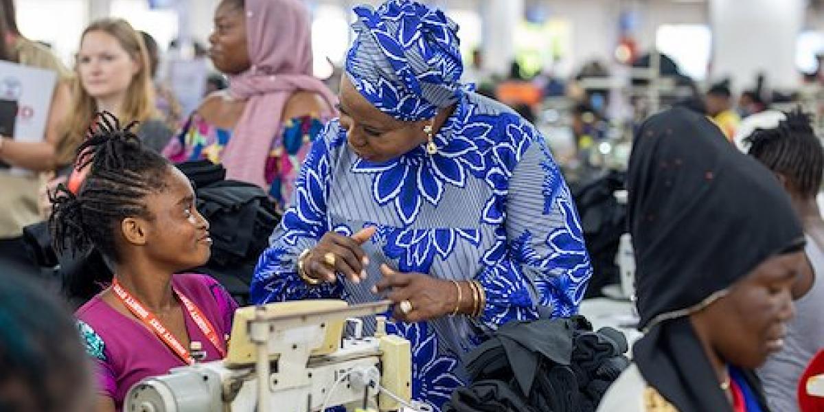 Woman in blue dress and headwrap speaks with woman sitting behind a sewing machine with other people in the factory behind them chatting with each other and working on sewing machines