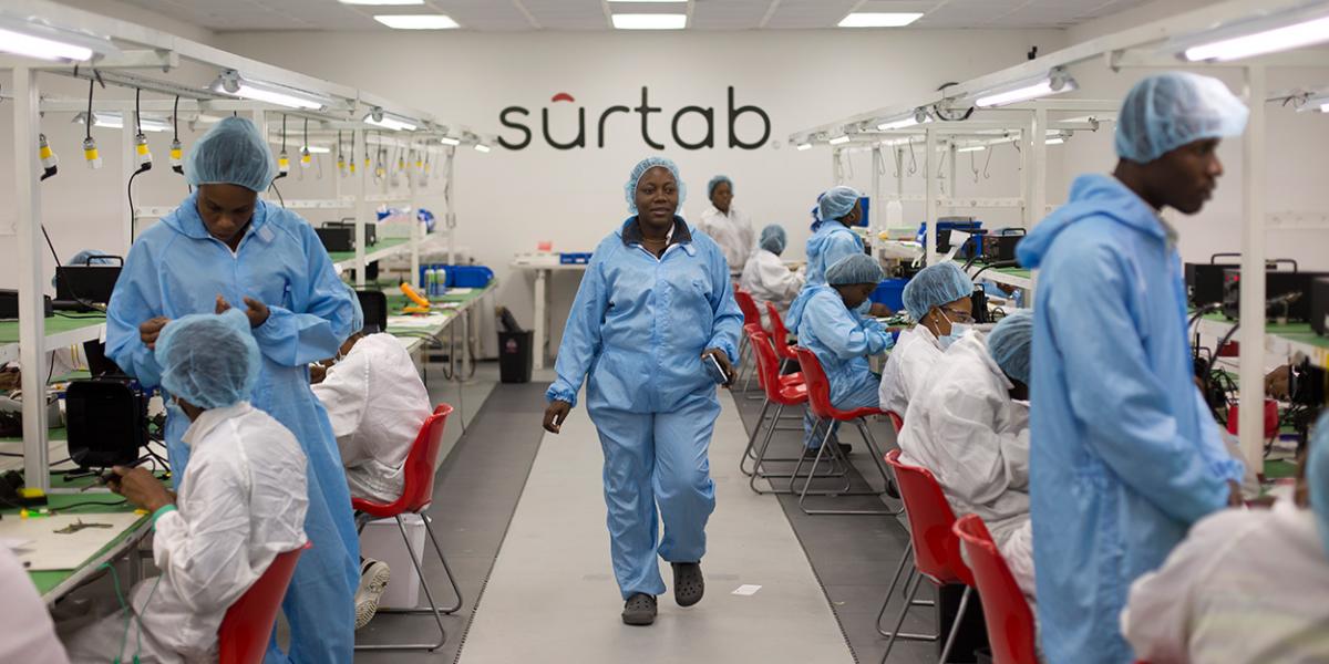 A woman in laboratory clothes walks between tables of workers assembling high-tech gadgets, with Surtab logo on the wall behind