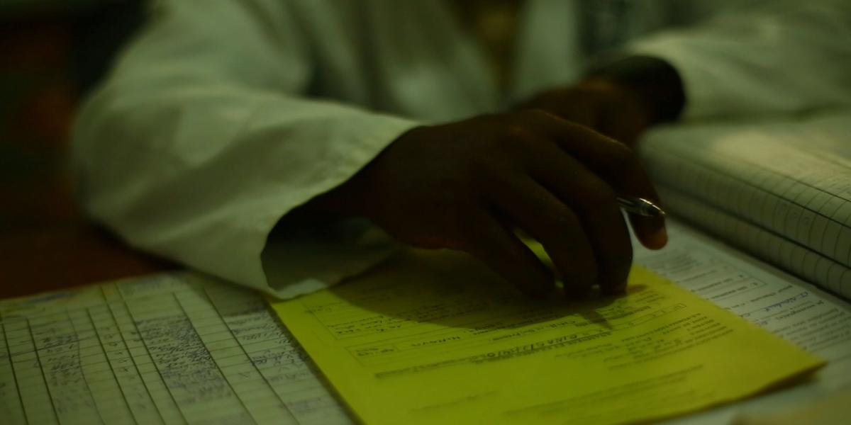 Closeup of a doctor's hands on a desk. He holds a pen and there are documents covering the desk.