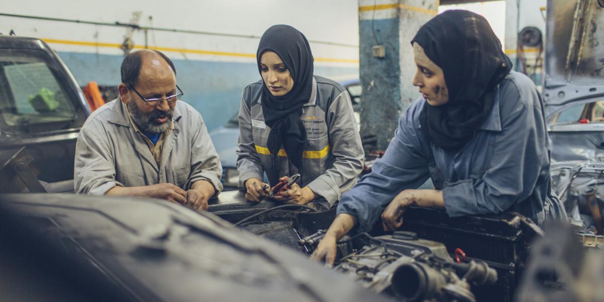 Najlae (left) and Rajae (right) look on as an instructor shows them how to repair part of a car.