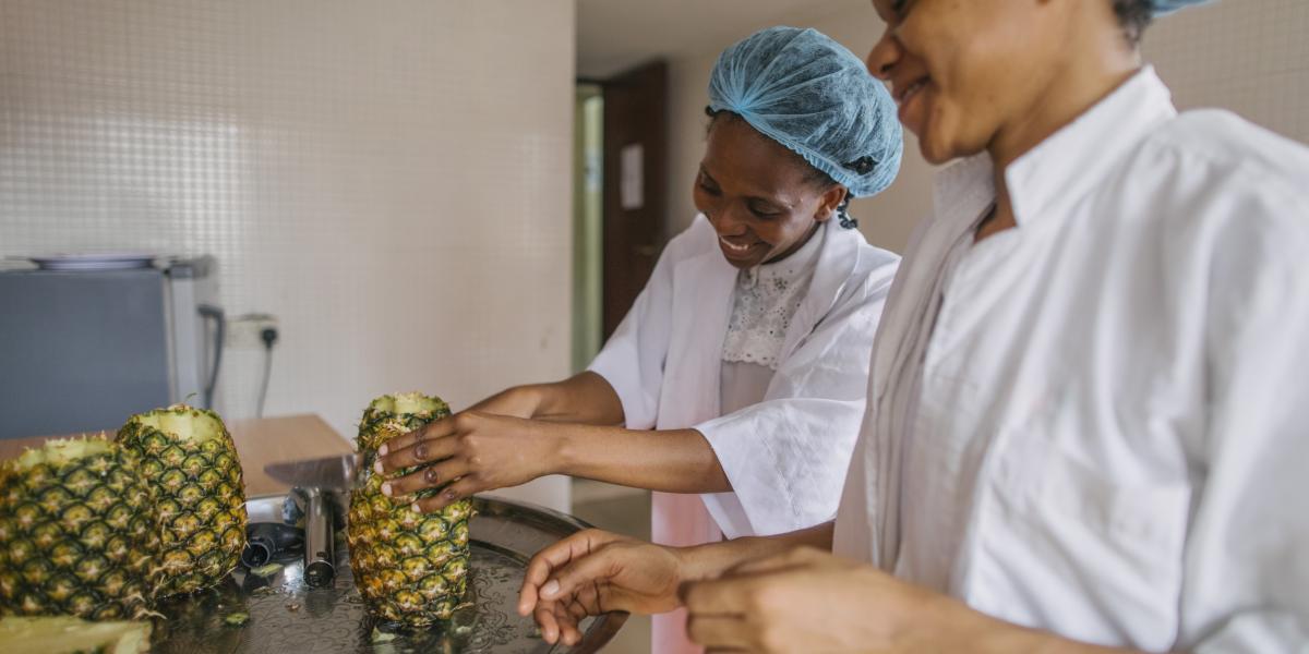 Two women cut pineapples in a kitchen.
