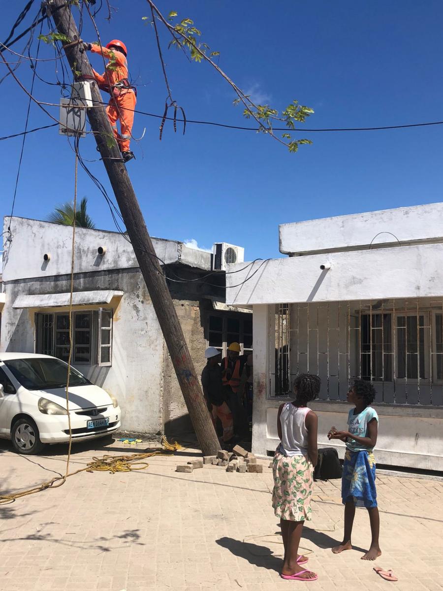 Two girls watch as Sidónia Figueira climbs a tilted pole to repair damage caused by Cyclone Gombe in March 2022