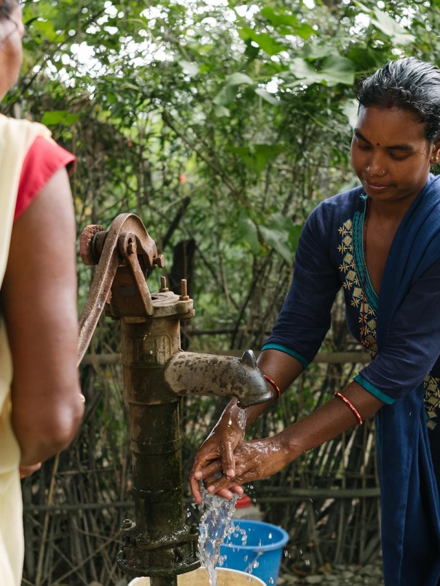 A woman washes her hands at an outdoor pump, while another woman pumps water