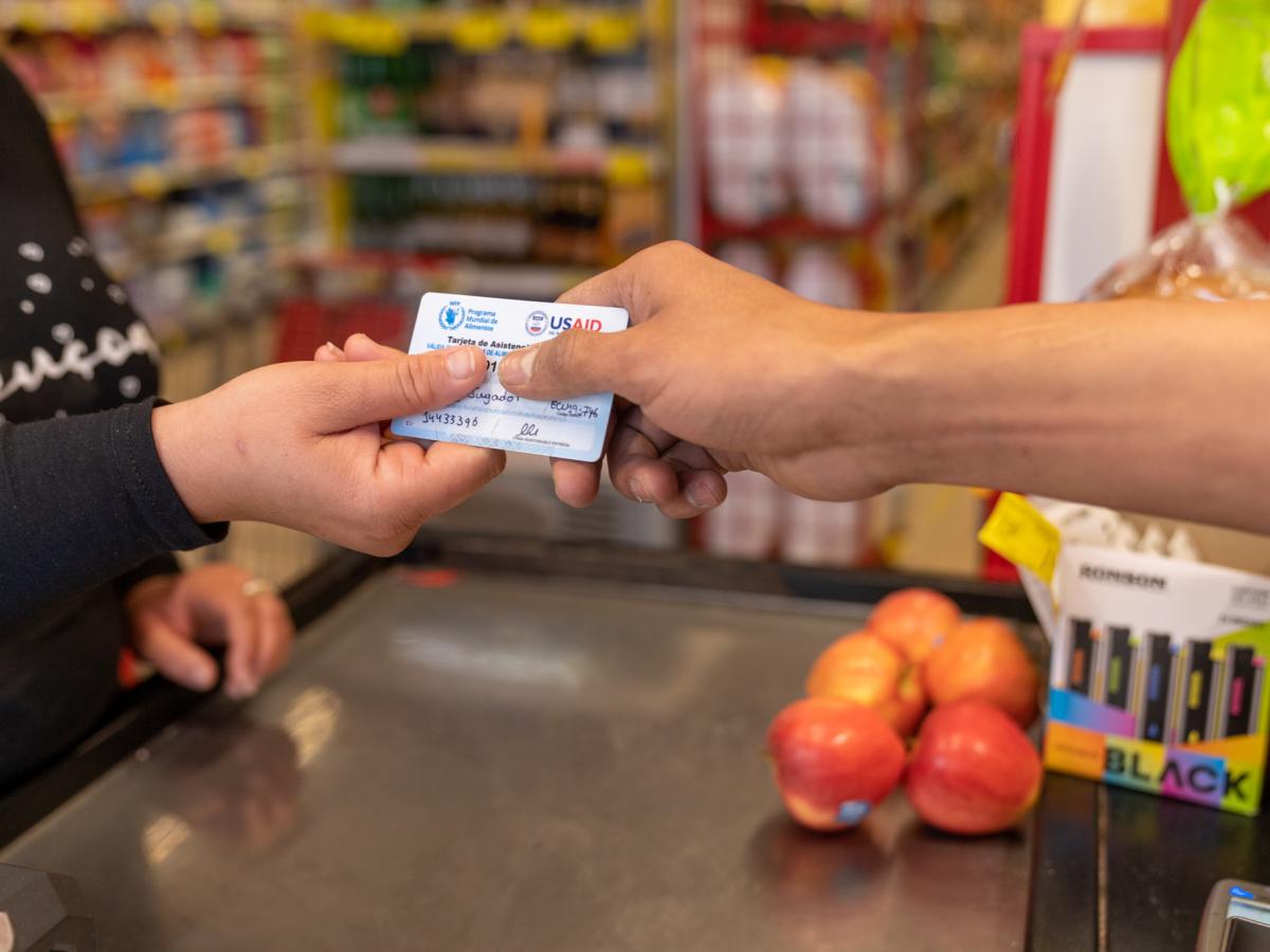 This picture shows a person handing a debit card to a cashier at a store. The debit card has the World Food Program and USAID logos.