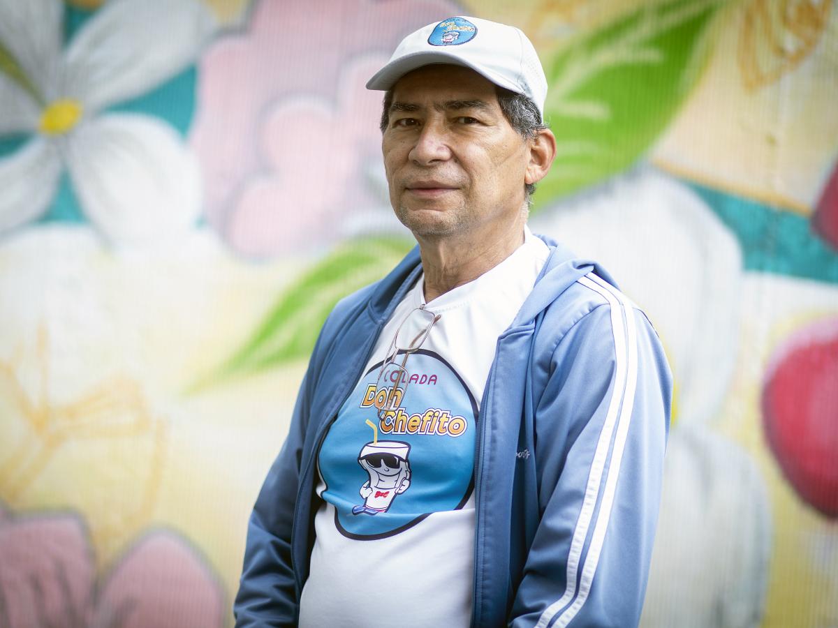 This picture shows Carlos Torres’ upper body and face. He is wearing a blue sports jacket and a hat, and is standing in front of a colorful mural with flowers.