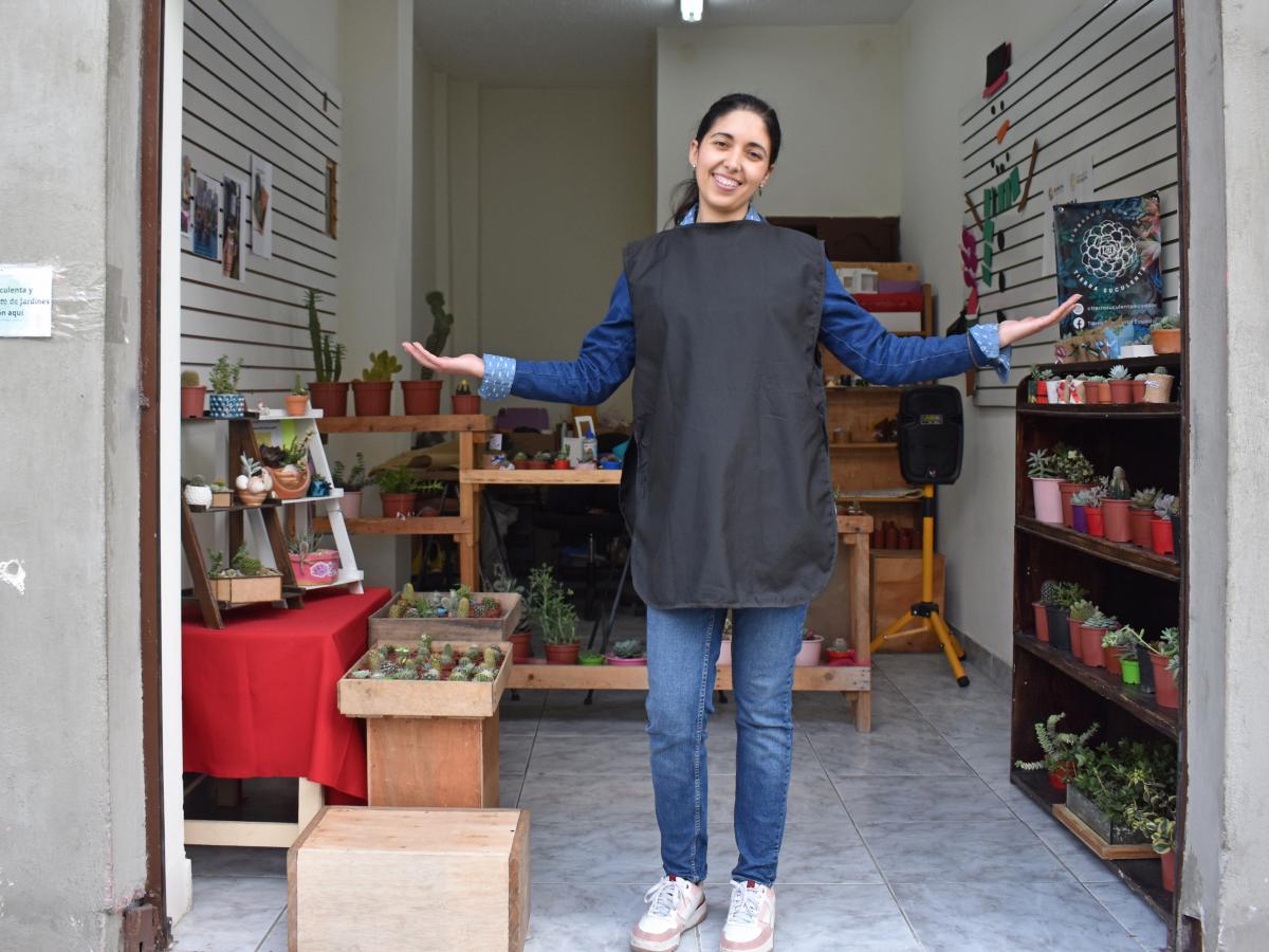 This is a picture of Omaira, the main character, standing in front of her little store with arms open, welcoming people. She is in jeans, a long-sleeved shirt, sneakers, and and black apron. There are plants on shelves.