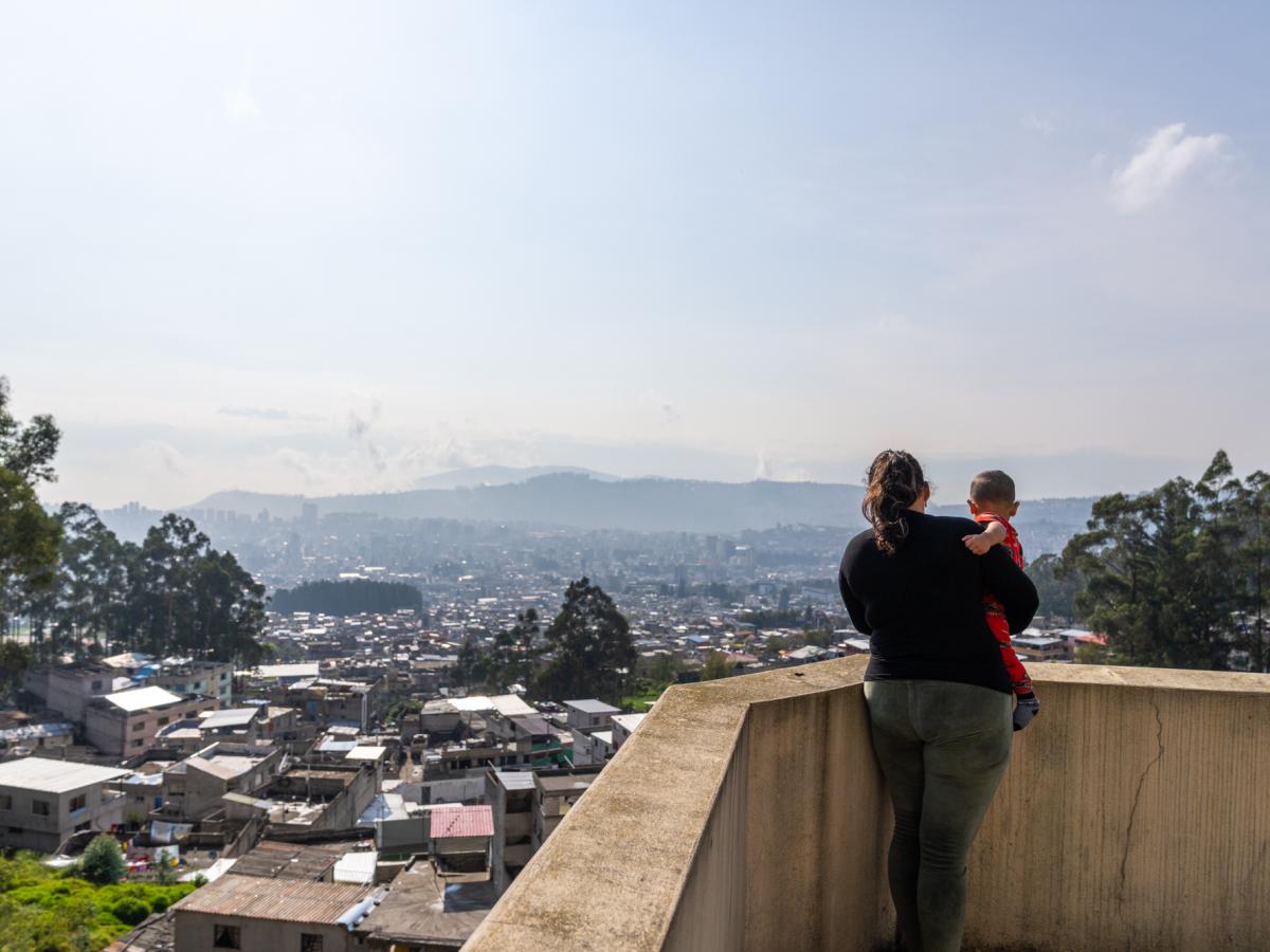 This picture shows Ariana, a female and main character in the story. She is standing on a balcony overseeing the city of Quito while holding her baby. The scenery shows buildings and trees in what is known to be the highlands of Ecuador. 
