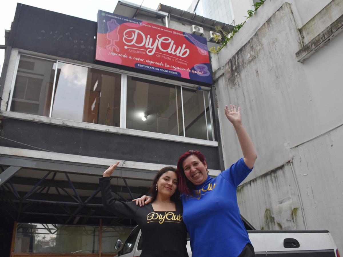 This picture shows aura and Dani standing in front of their building. Both are raising their hands in excitement. There is a sign on the building that reads DIY Club. 