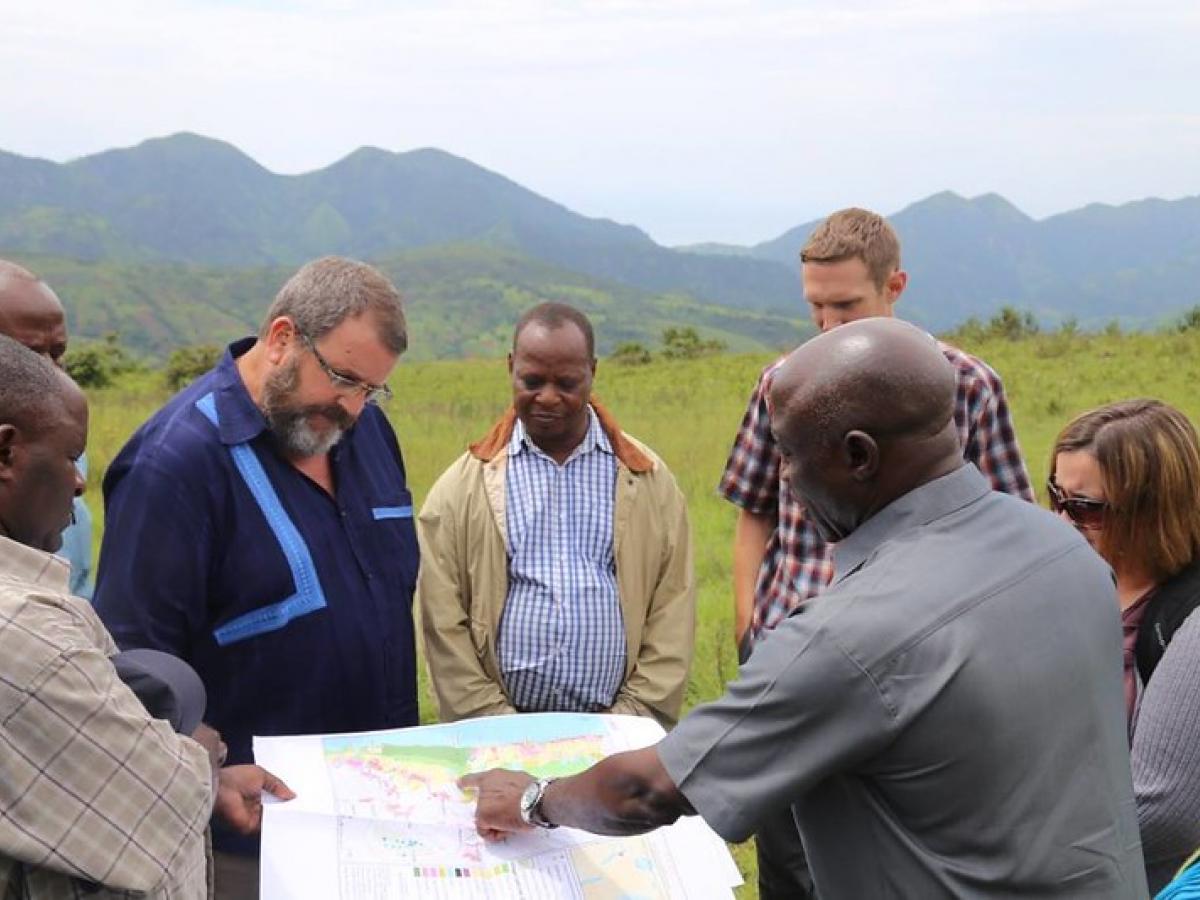 A group of people in front of a mountainous landscape reviewing a large printed map (Photo Credit: USAID Tanzania)
