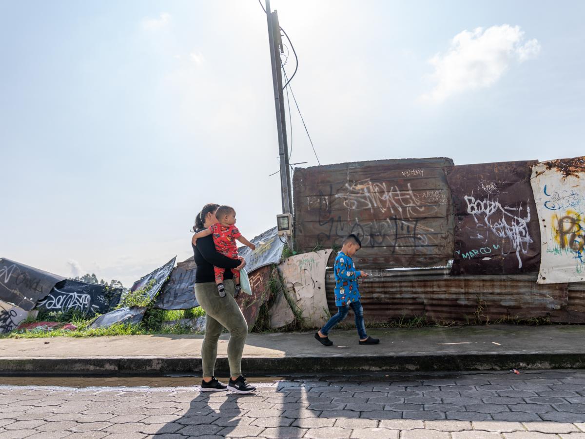This picture shows Ariana, a female and main character in the story, and her two boys walking on the street. There is a wall with scrap metal sheets and graffiti on it.