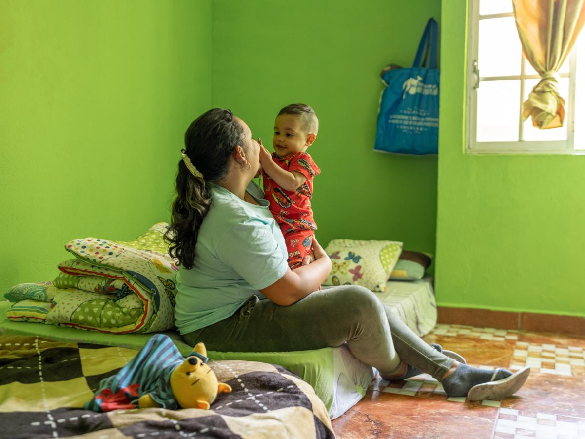 This picture shows Ariana, a female and main character in the story, and her one-year old sitting on a mattress on the floor. The baby is playing with her face and smiling. They are in a green room with a window and sunlight coming in. 