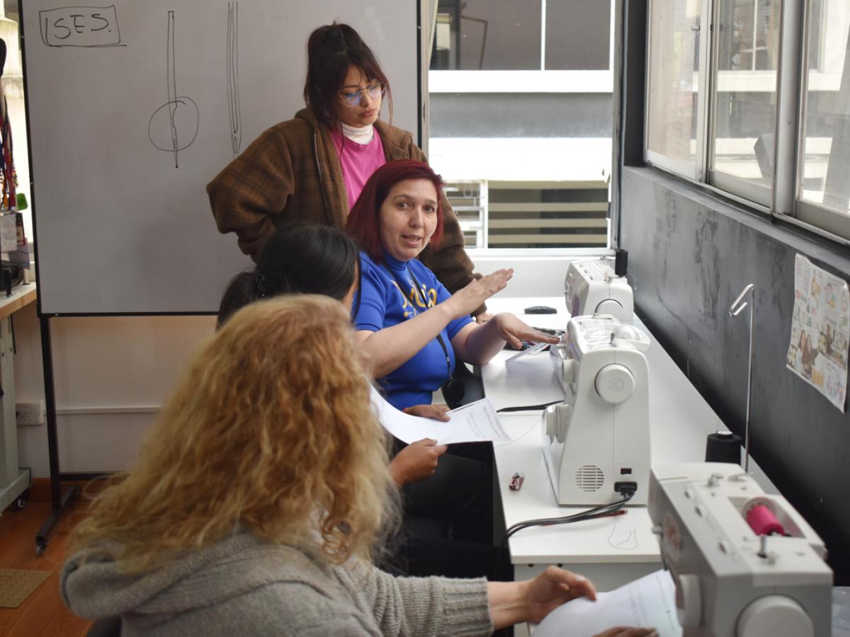 This picture shows Laura teaching three female students how to use a sewing machine. The students are all paying close attention to Laura. 