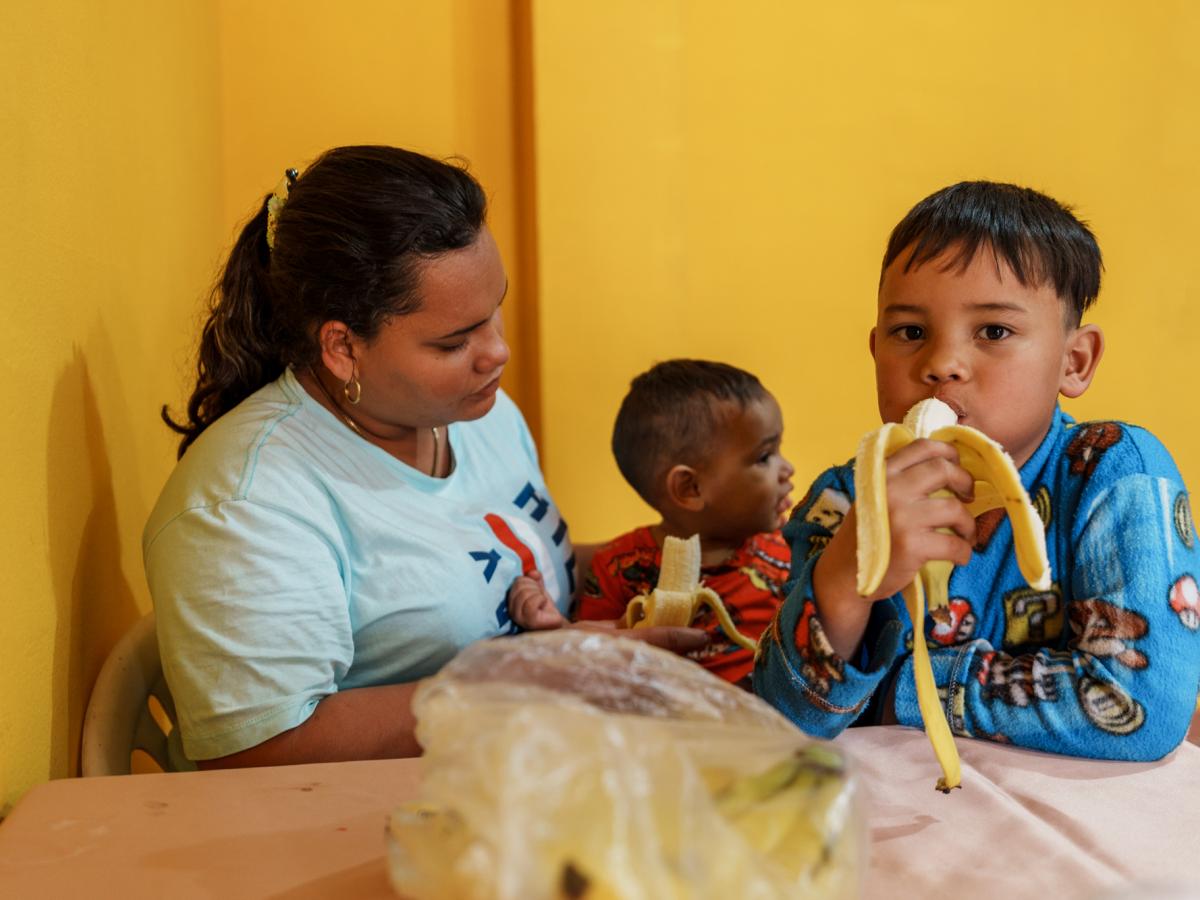 This picture shows Ariana, a female and main character in the story, with her two boys. They are sitting at a table in a yellow-colored room and eating bananas. 