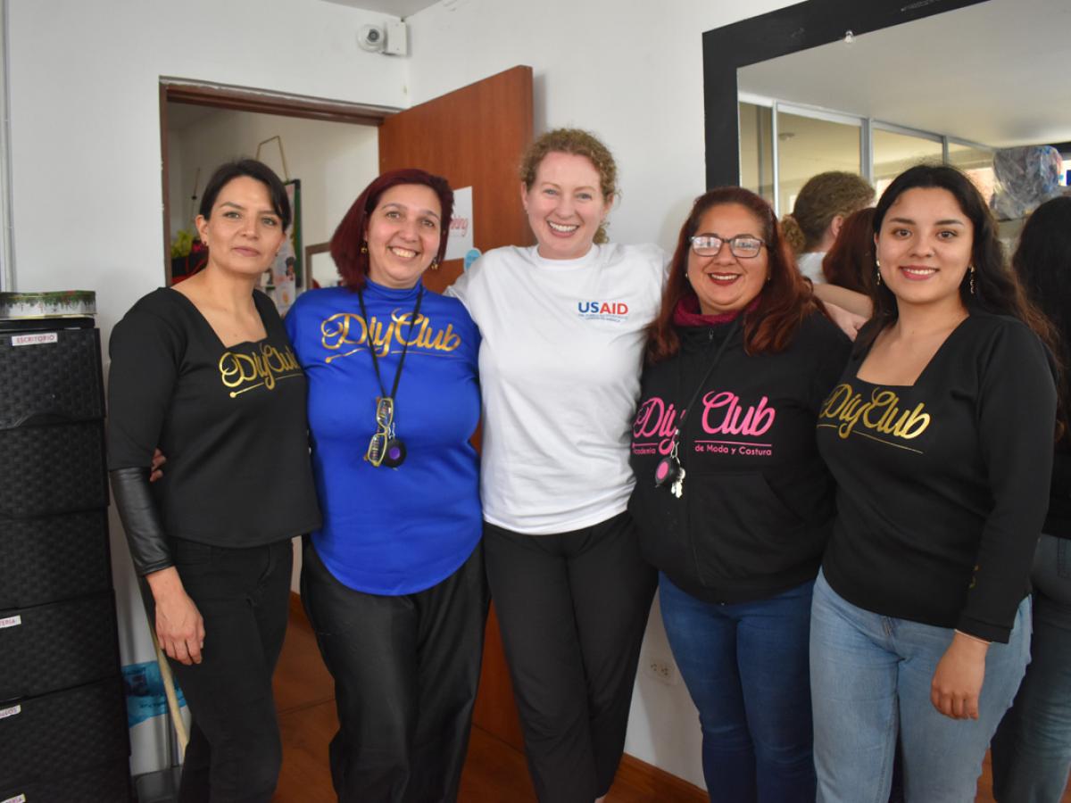 This picture shows five females all standing in a row next to each other. Two of them are Laura and Dani, the other two are hired members of the staff, and the fifth lady is a USAID employee. They are all smiling and wearing shirts with their organization’s identities. 