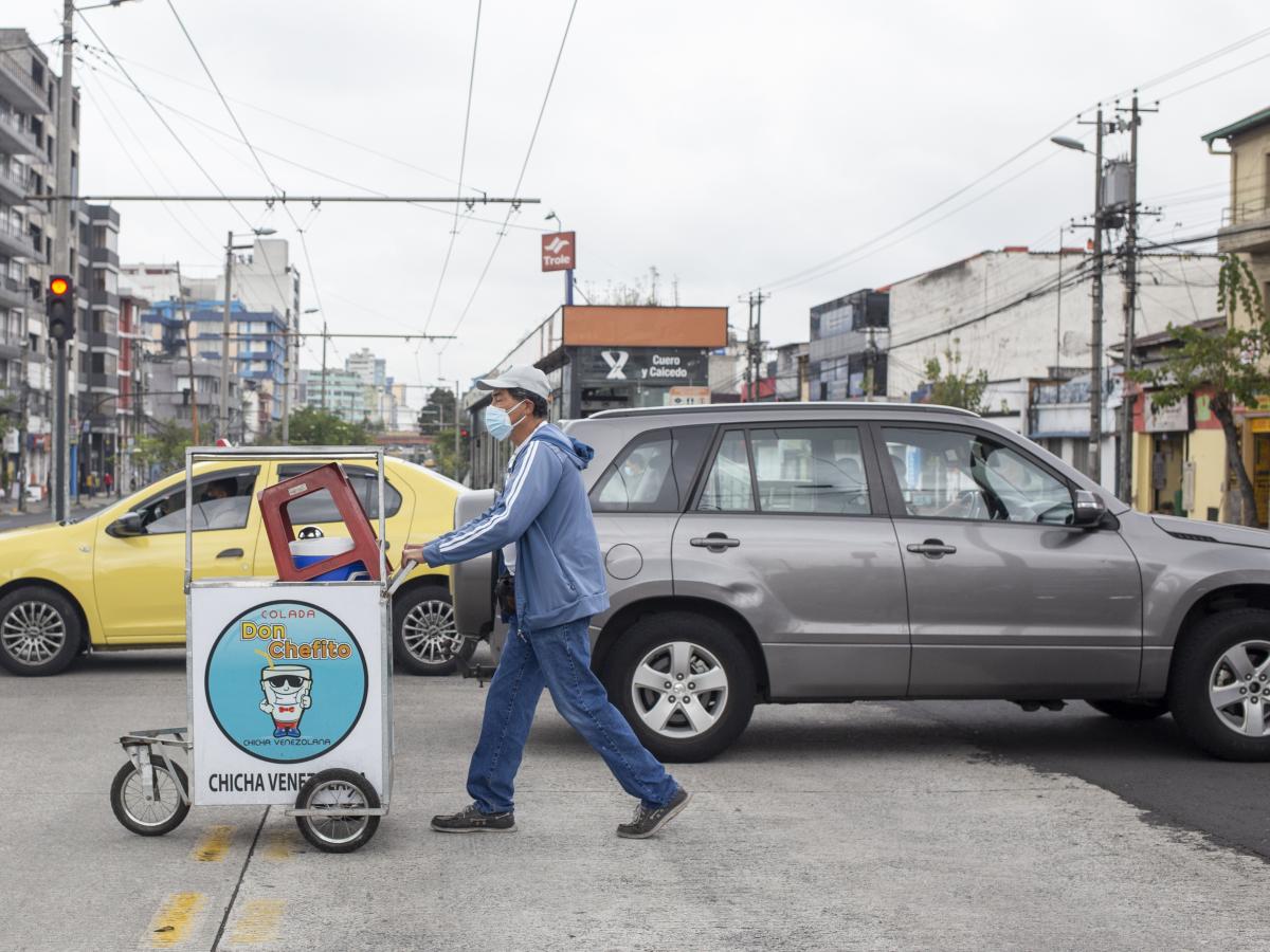 This picture shows Carlos Torres pushing his vendor cart on the streets of Quito. Other vehicles are also passing by. There are buildings in the background. 