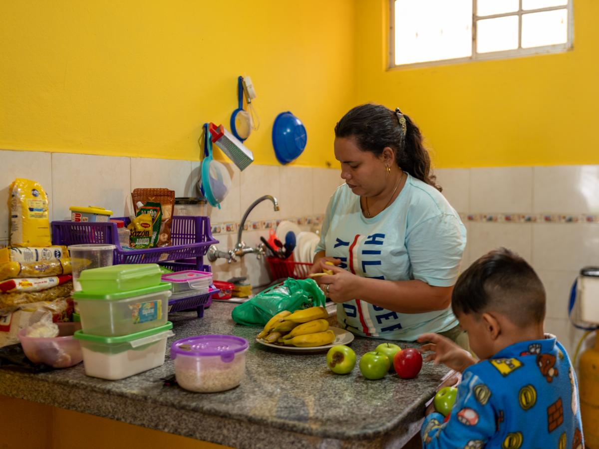 This picture shows Ariana, a female and main character in the story, with her three-year old son putting groceries away. They are in the kitchen. Groceries include fruits such as bananas and apples. 