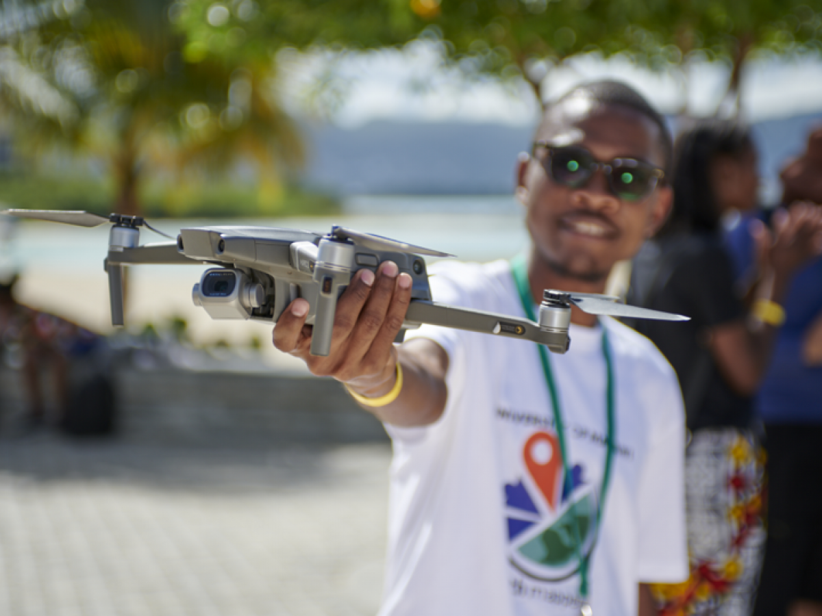 Photo of a young man in a YouthMappers t-shirt holding up a drone (Photo Credit: YouthMappers)
