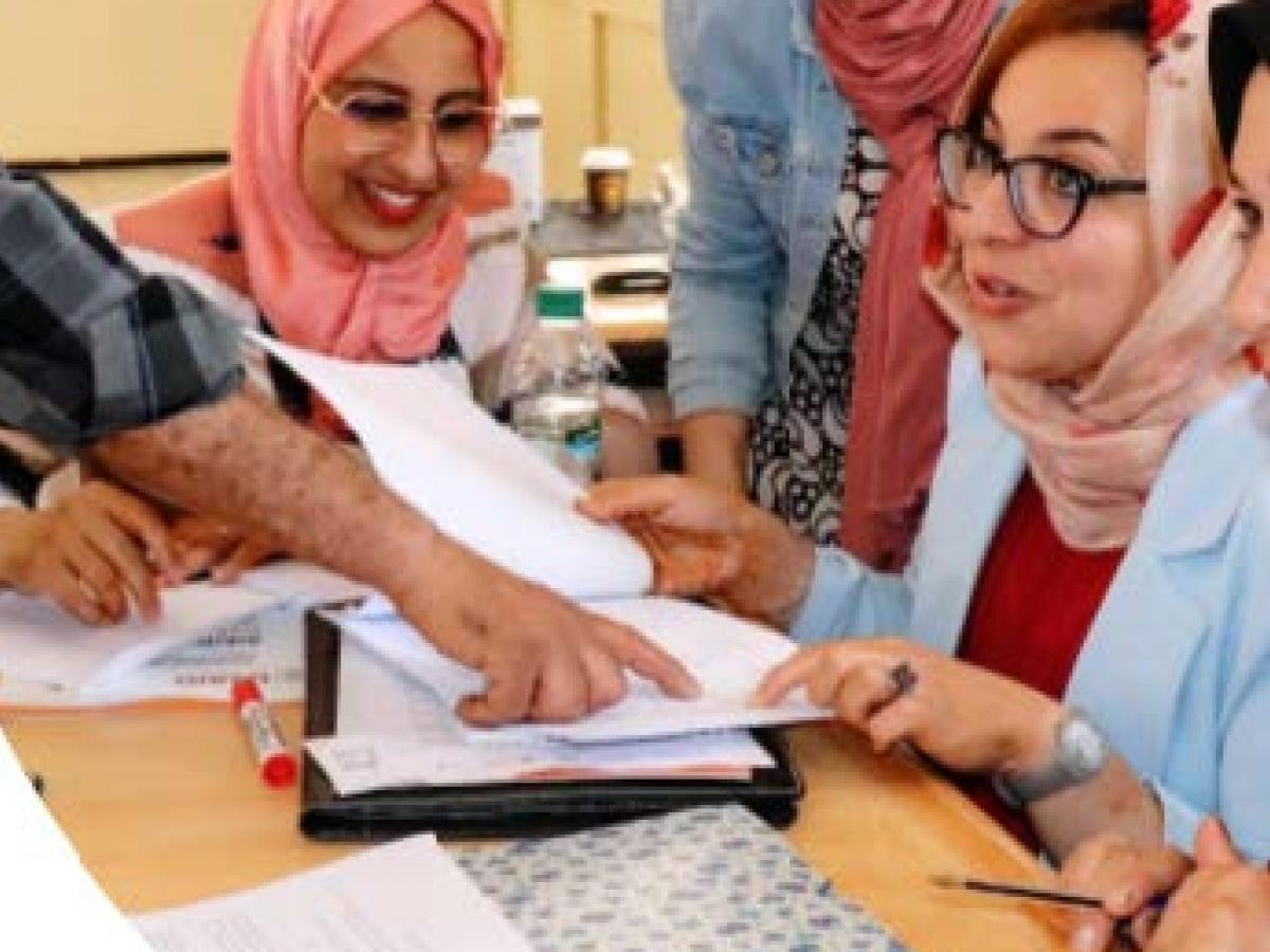 A few women wearing hijab interact as a man who is out-of-frame points to something written on a piece of paper on the desk where they are seated.