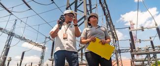 Engineers standing outside a power transmission facility. High-power energy transmission lines appear above them.