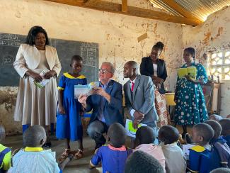 Ambassador Young kneels next to a young student in a classroom, holding a children's picture book that was printed by USAID.
