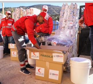 A young man dressed in Red Crescent uniform pulls a bowl out of box marked "fragile" and stickers portraying the logos of USAID and the Moroccan Red Crescent. Other Red Crescent volunteers observe. Packages of light mattresses and a round mountain peak appear in the background.