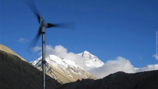 Photo of a wind turbine against a background of mountaintops, puffy clouds, and a blue sky.