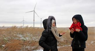 Two happy women in winter clothes chat in a dry amber field with wind turbines in the distance set against a gray sky