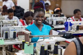 Smiling woman sitting behind a sewing machine with rows of women and men work behind sewing machines